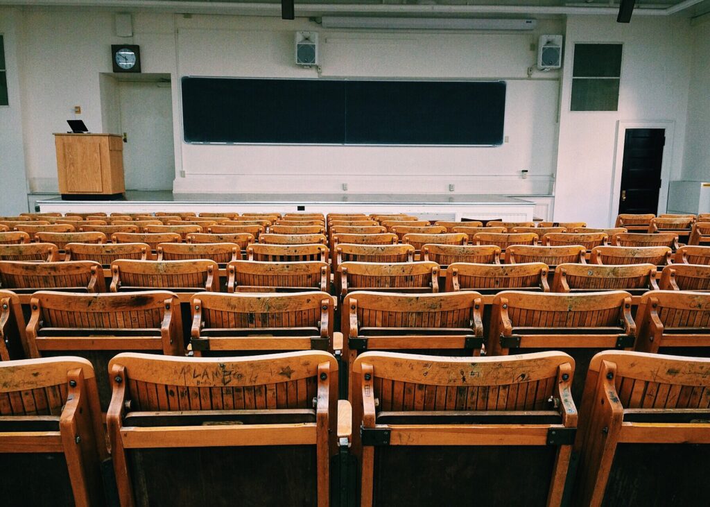 Hier ist ein leerer Hörsaal abgebildet. Der Betrachtende steht weiter oben in einem Hörsaal und blickt über leere, hellbraune Stühle in Richtung einer Tafel. Mit diesem Bild ist die Seite "Good Practice aus der Hochschullandschaft" verlinkt.