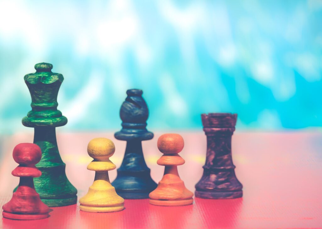 Auf diesem Bild stehen sechs unterschiedliche Schachfiguren in verschiedenen Farben auf einem roten Tisch. Sie stehen in zwei Reihen versetzt. Mit diesem Bild ist die Seite "Lehrendenprofile für Gamification" verlinkt.