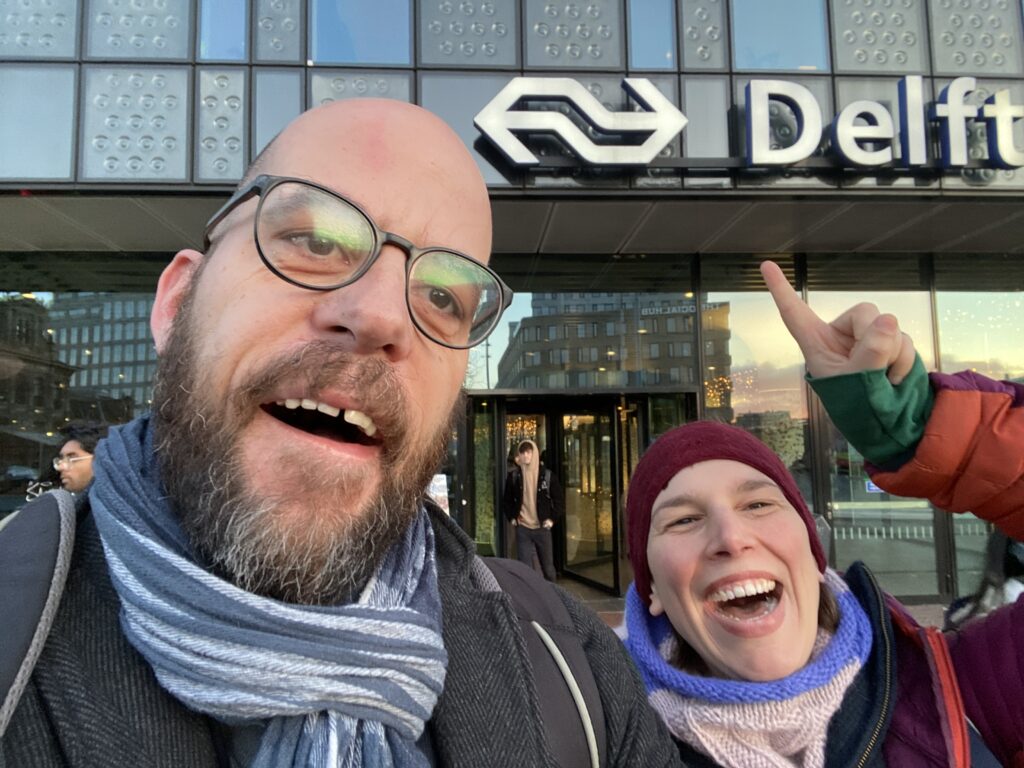 Markus Deimann und Paulina Rinne bei der Ankunft am Delfter Hauptbahnhof. Beide blicken glücklich und aufgeregt in die Kamera. Paulina Rinne deutet auf die Aufschrift "Delft", die über ihnen am Bahnhof prangt. 
