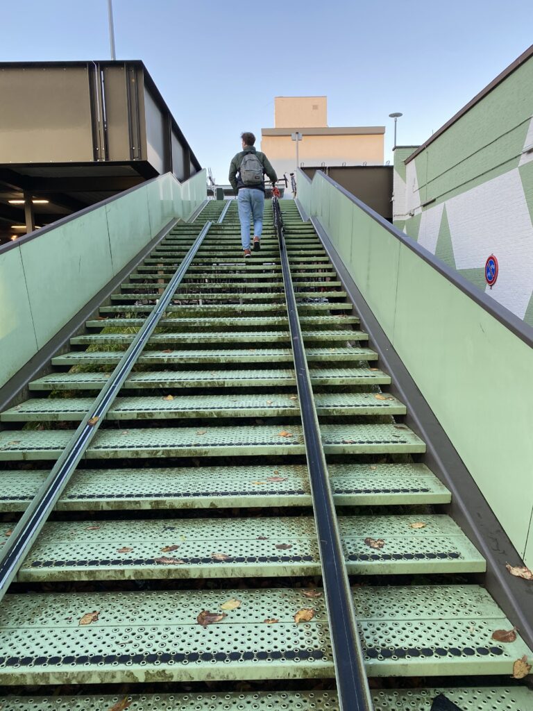 Ein Fahrradfahrer der sein Fahrrad eine Fahrradschiene hinaufschiebt. Die Treppe, das Geländer sowie die Stufen sind Türkis. Der Himmel im Hintergrund ist Hellblau. 