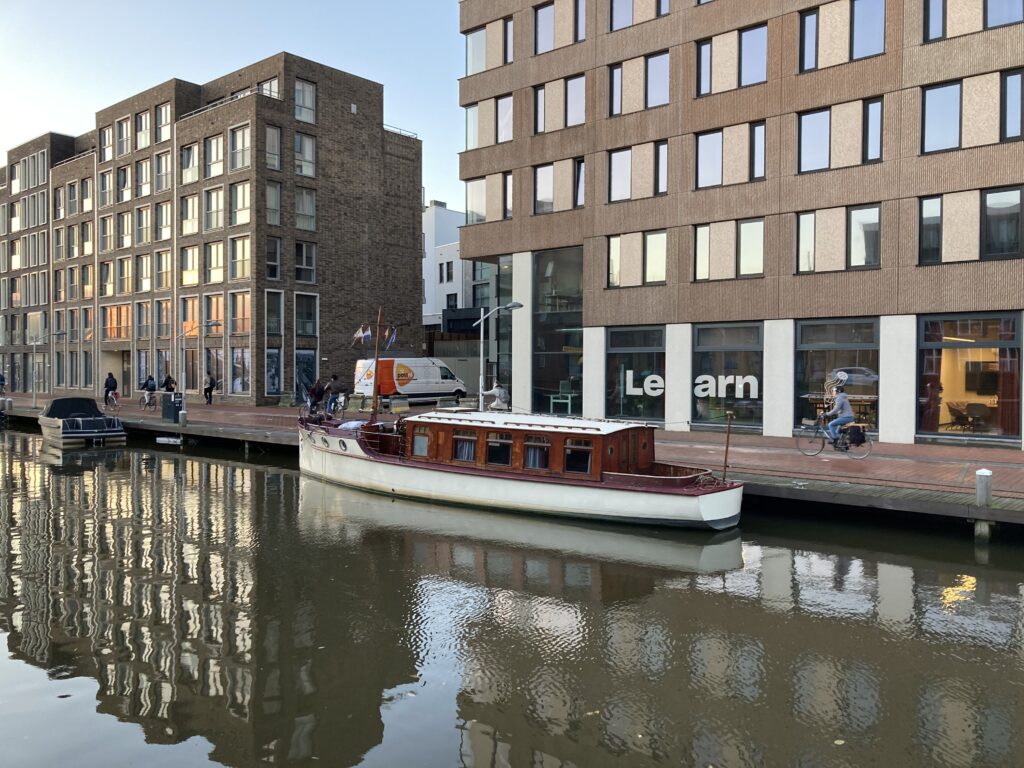 Im Vordergrund eine für Delft typische Gracht, darauf ein kleines, rustikales Boot. Im Hintergrund einige Fahrradfahrer und ein hohes Gebäude. In diesem Gebäude unten auf der Fensterfront die Aufschrift in großen Buchstaben: Learn. 