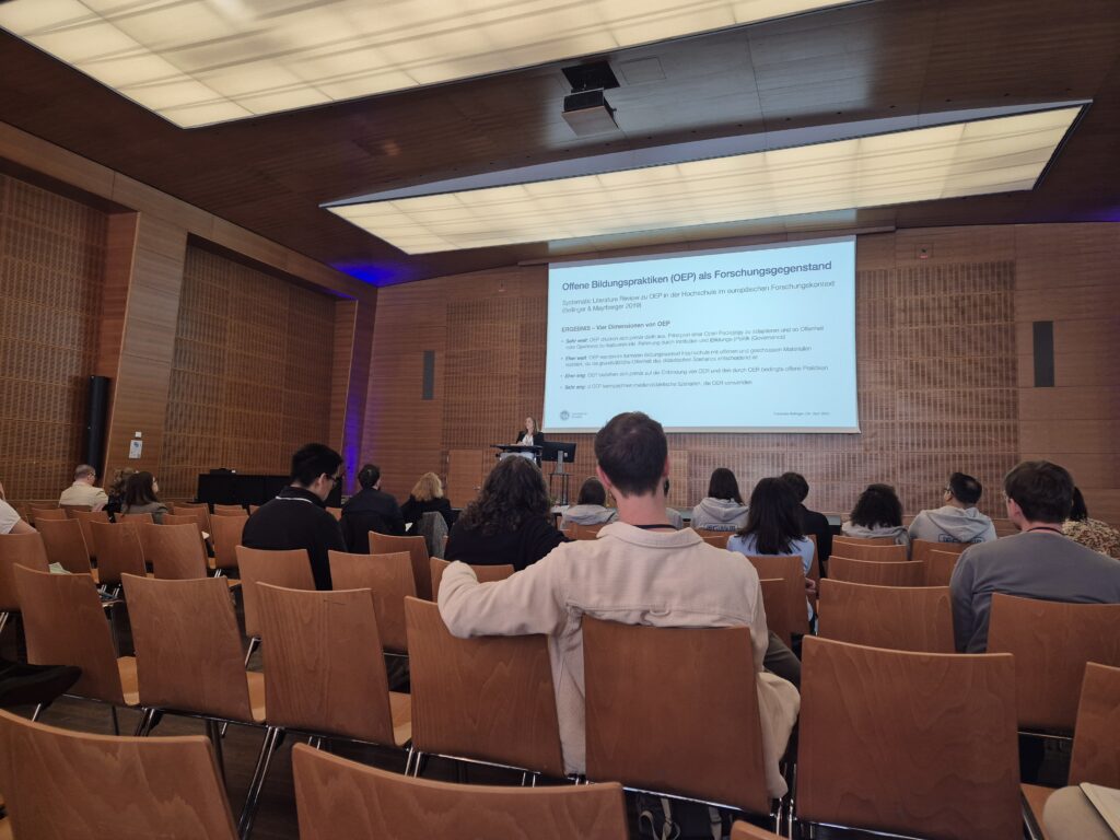 Einige Menschen sitzen in einem Konferenzsaal in Stuhlreihen und schauen zu einer Bühne. Auf der Bühne steht eine Person am Pult.