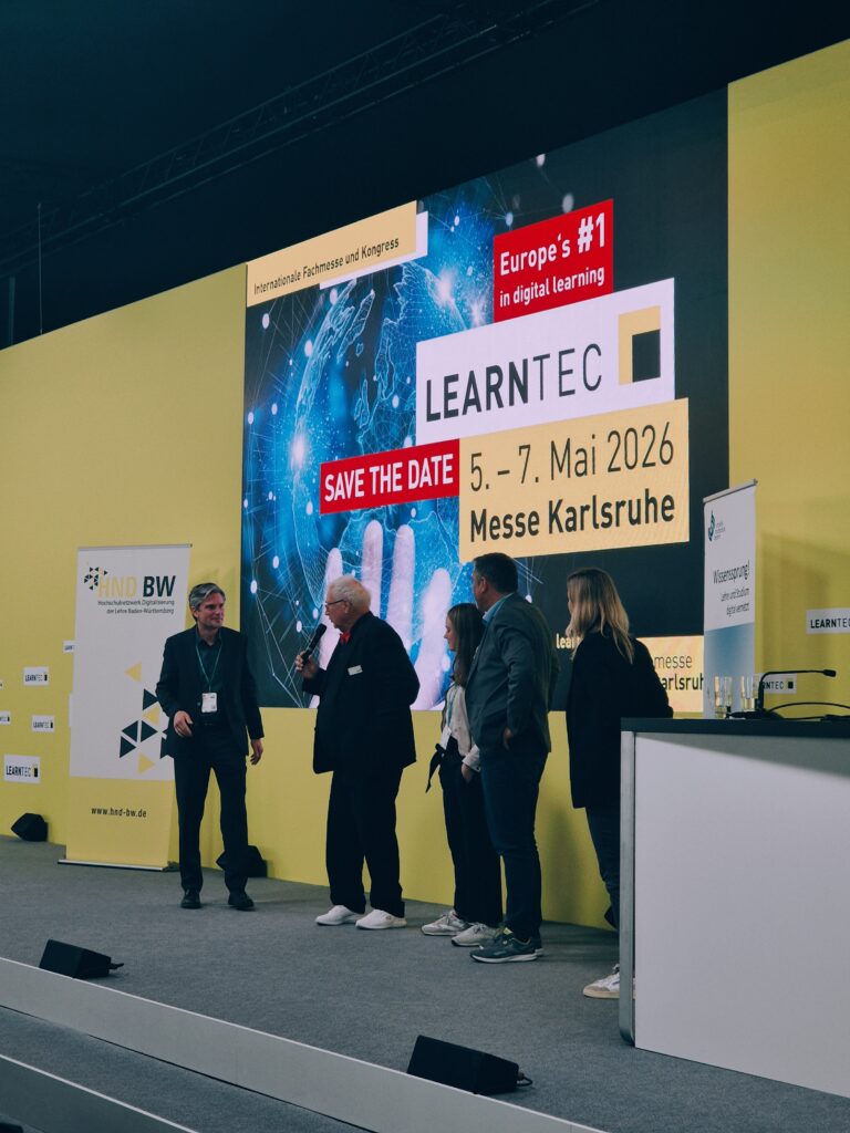 Fünf Personen stehen auf einer Bühne. Eine Person hält ein Mikrofon . Dahinter steht auf einem projizierten Bild "LEARNTEC".