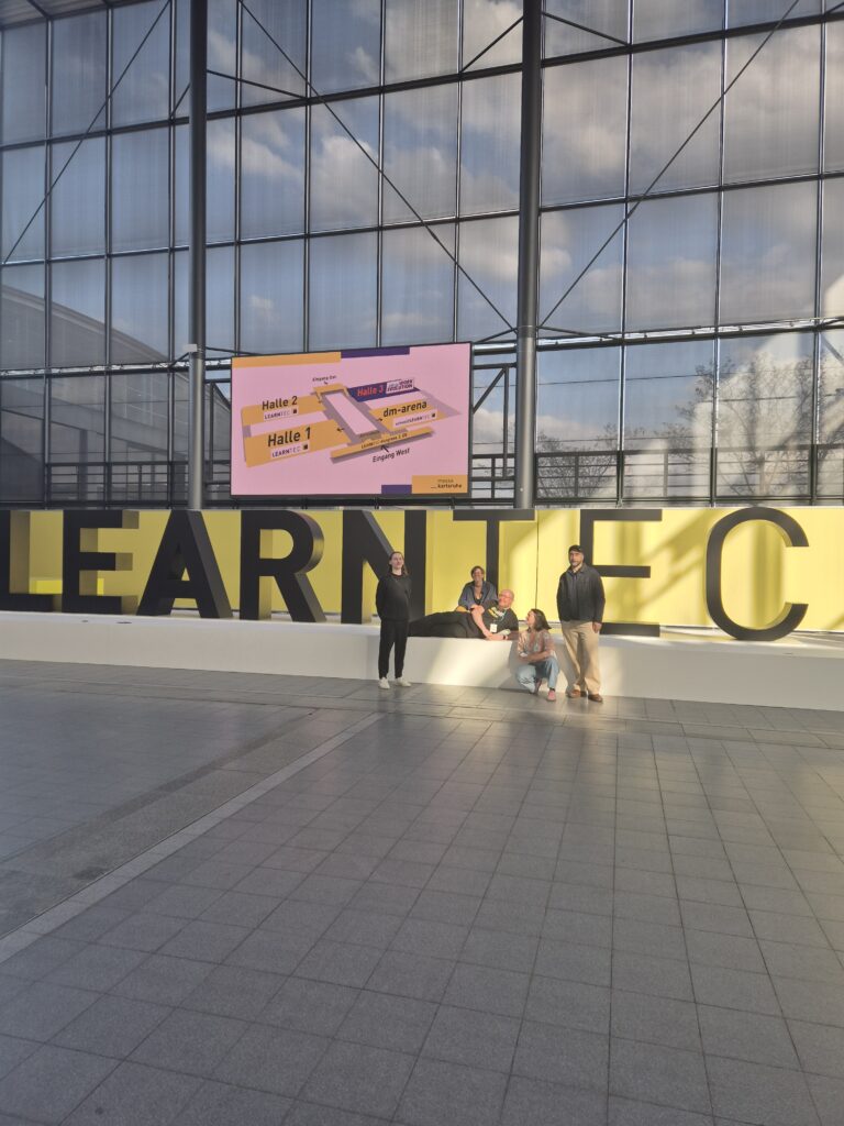 Ein großer Schriftzug "LEARNTEC" auf eine Podest in einer Halle. Davor stehen und sitzen fünf Menschen.