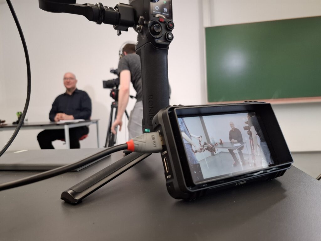 Über einen kleinen Bildschirm im Vordergrund ist zu sehen, was durch die Videokamera aufgenommen wird. Im Hintergrund ist unscharf zu erkennen, dass eine Person am Tisch sitzt. Daneben ein Kameramann.