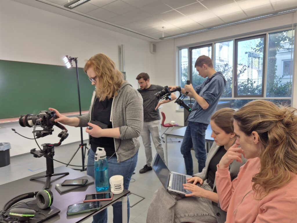 Hinter den Kulissen der Videoaufnahmen sind fünf Personen an Laptop und Kameras beschäftigt.