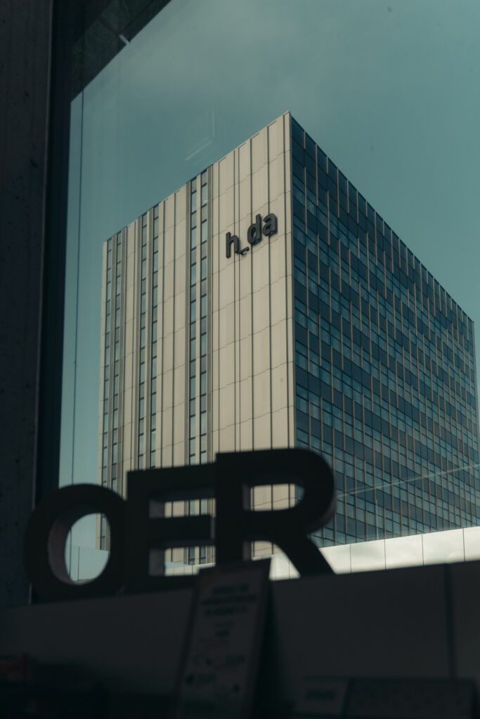 Blick durch ein Fenster auf ein Hochhaus der Hochschule Darmstadt (h_da); im Vordergrund unscharf die Buchstaben OER.