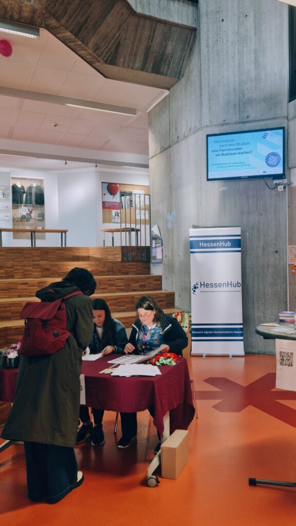 Zwei Menschen sitzen an einem Tisch in einem Foyer und verteilen Namensschilder. Davor steht eine weitere Person. Im Hintergrund steht ein Aufsteller mit dem Schriftzug "HessenHub".  