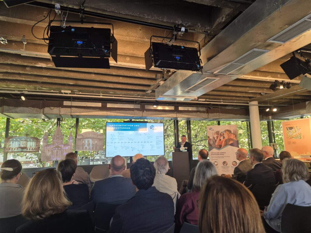 Foto: Eine Person steht hinter einem Pult und neben einem Bildschirm. Dort ist ein Zeitstrahl zu sehen. Davor sitzen Personen in Sitzreihen und blicken zum Redner.