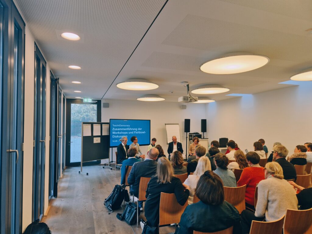 Personen sitzen in mehreren Reihen in einem Seminarraum und blicken nach vorn. Vorne sitzen 5 Personen in einem Halbkreis vor einem Bildschirm. Auf de Bildschirm steht "Zusammenführung der Ergebnisse und Fishbowl-Diskussion".