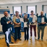 Sechs Personen stehen nebeneinander und lächeln in die Kamera. Vier Personen in der Mitte halten eine Urkunde und einen Preis in Form eines Löwens. Die Person links hält einen Bembel-Krug in der Hand.