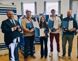 Sechs Personen stehen nebeneinander und lächeln in die Kamera. Vier Personen in der Mitte halten eine Urkunde und einen Preis in Form eines Löwens. Die Person links hält einen Bembel-Krug in der Hand.