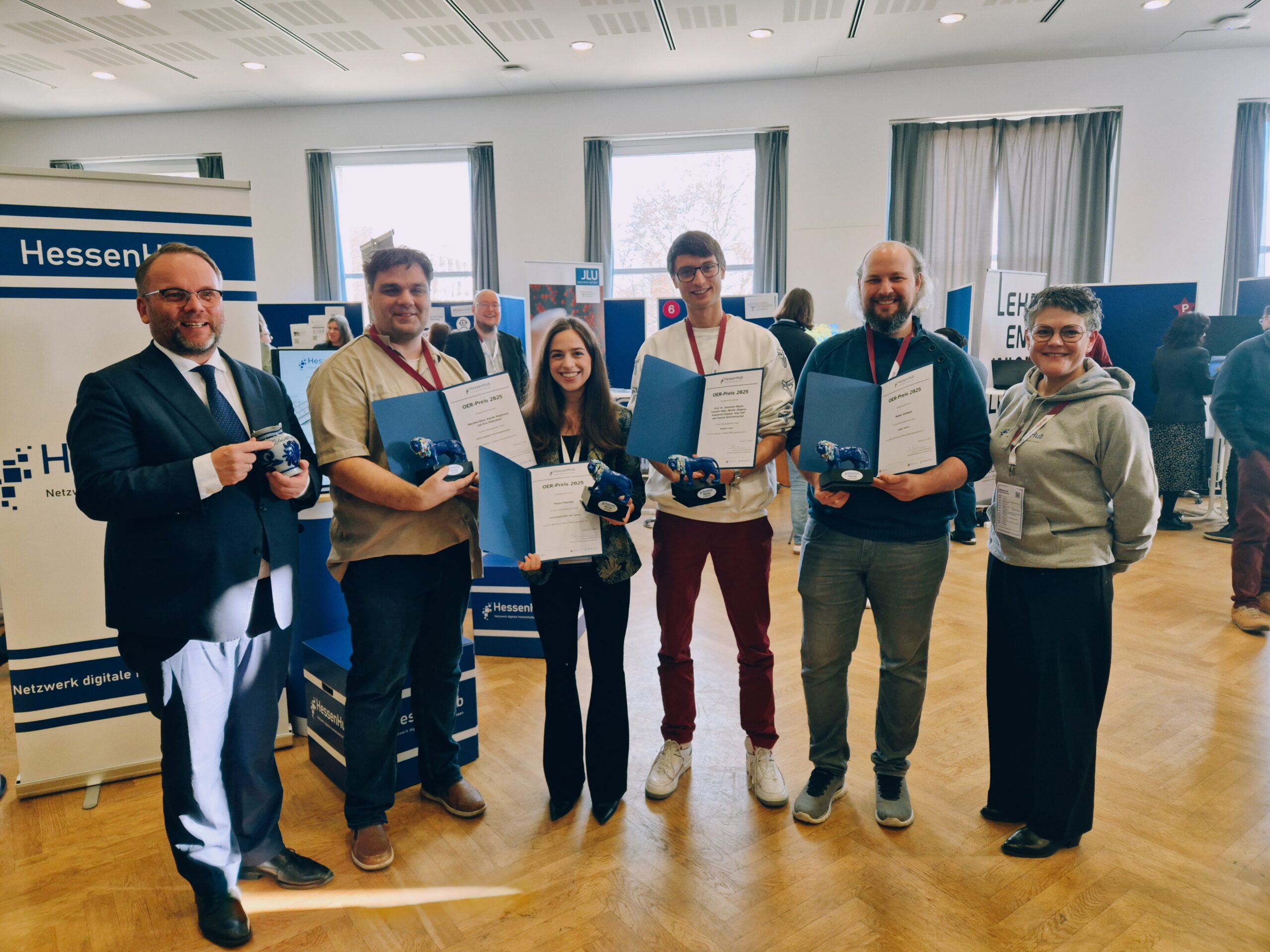 Gruppenfoto der OER-Preisträger*innen mit Vizepräsidenton Yvonne Zimmermann und Wissenschaftsminister Timon Gremmels bei einer Veranstaltung: Mehrere Teilnehmende halten Urkunden und kleine blaue Preise in den Händen und stehen in einem hellen Veranstaltungsraum mit Informationsständen im Hintergrund