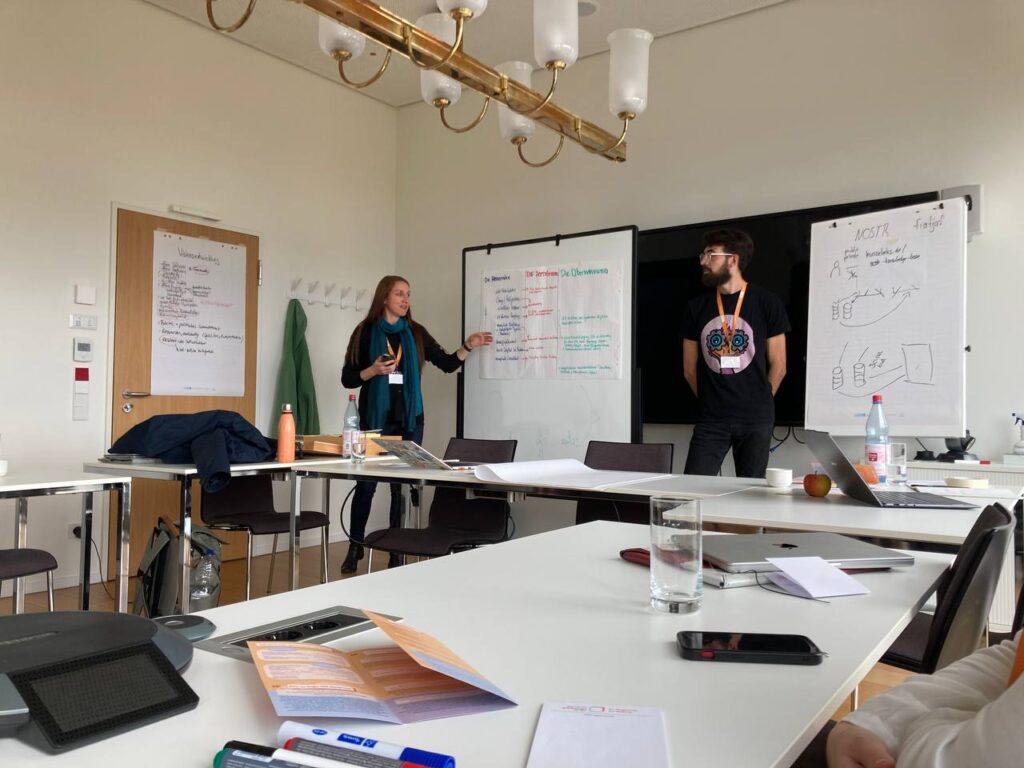 Foto: Zwei Personen stehen in einem Seminarraum vor mehrere Flipcharts und selbst gemalten Postern.