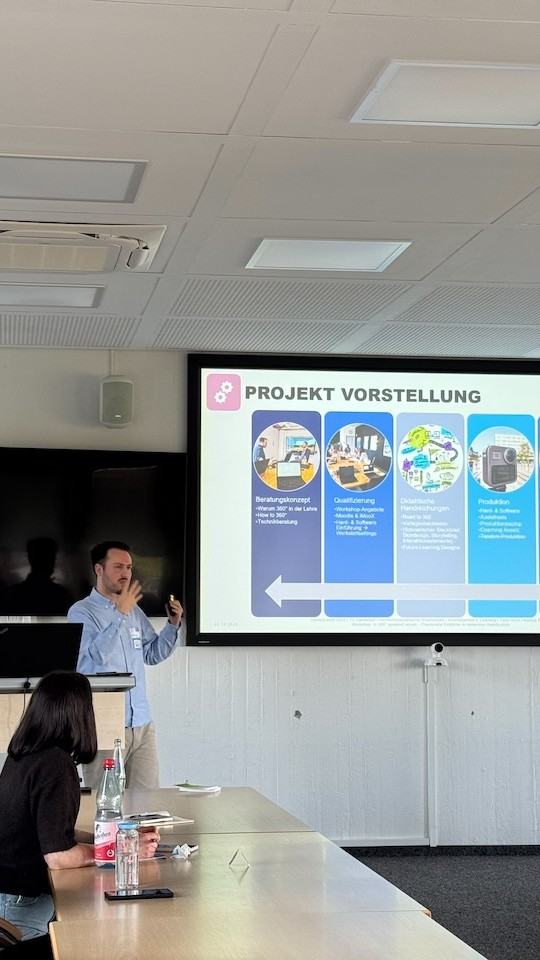 Eine Person steht in einem Seminarraum vor einer Leinwand. Auf diese wird eine Folie projiziert. Dort steht "Projektvorstellung". Davor sitzt eine Person an einem Tisch und  blickt die referierende Person an.