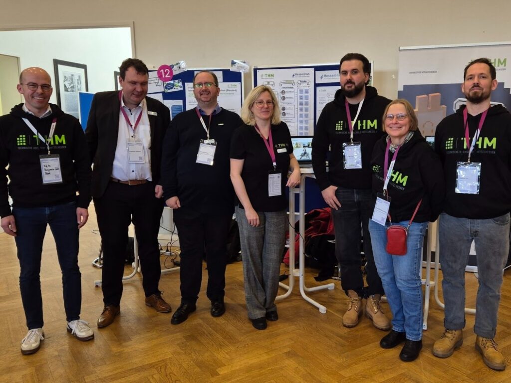 Foto von sieben Kolleg*innen der THM in schwarzen Pullovern mit grünem THM-Logo vor ihrem Stand während der EduConnect