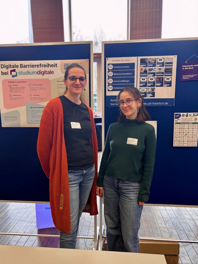 Vor zwei mit Postern und Material behängten Stellwänden stehen zwei weibliche Personen (Sanja Grimminger und studentische Hilfskraft Alexandra) und lächeln in die Kamera. 
